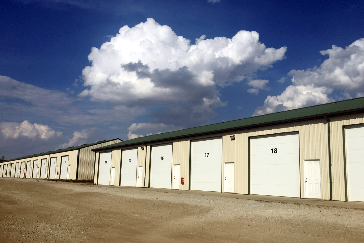 Two storage unit buildings with gravel leading to concrete approach, green roofs, tall white overhead garage doors that are numbered, and white entry doors with deadbolts.