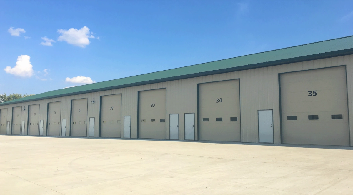 A storage unit building with paved approach, green roof, tall overhead garage doors that are numbered with three small windows in each, and white entry doors with deadbolts.